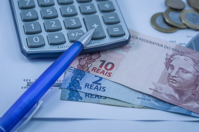 Imagem mostra calculadora, caneta, cédulas e moedas em cima de papéis